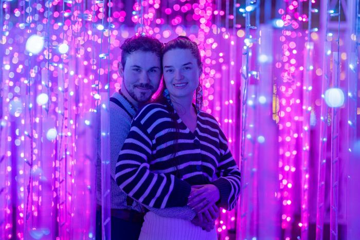 Ava and Bran standing close in a curtain of purple and pink lights